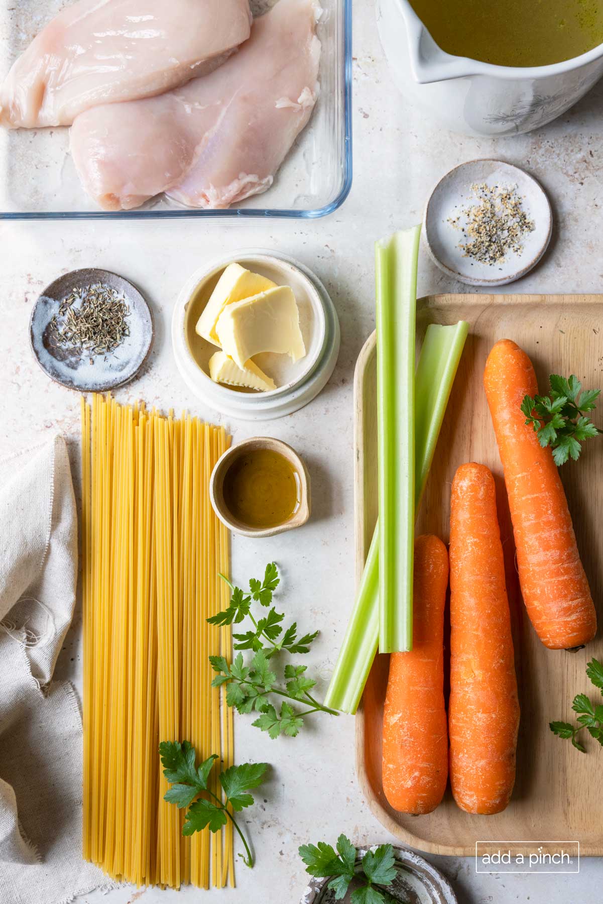 Chicken, broth, seasonings, butter, vegetables, and pasta on a white surface.