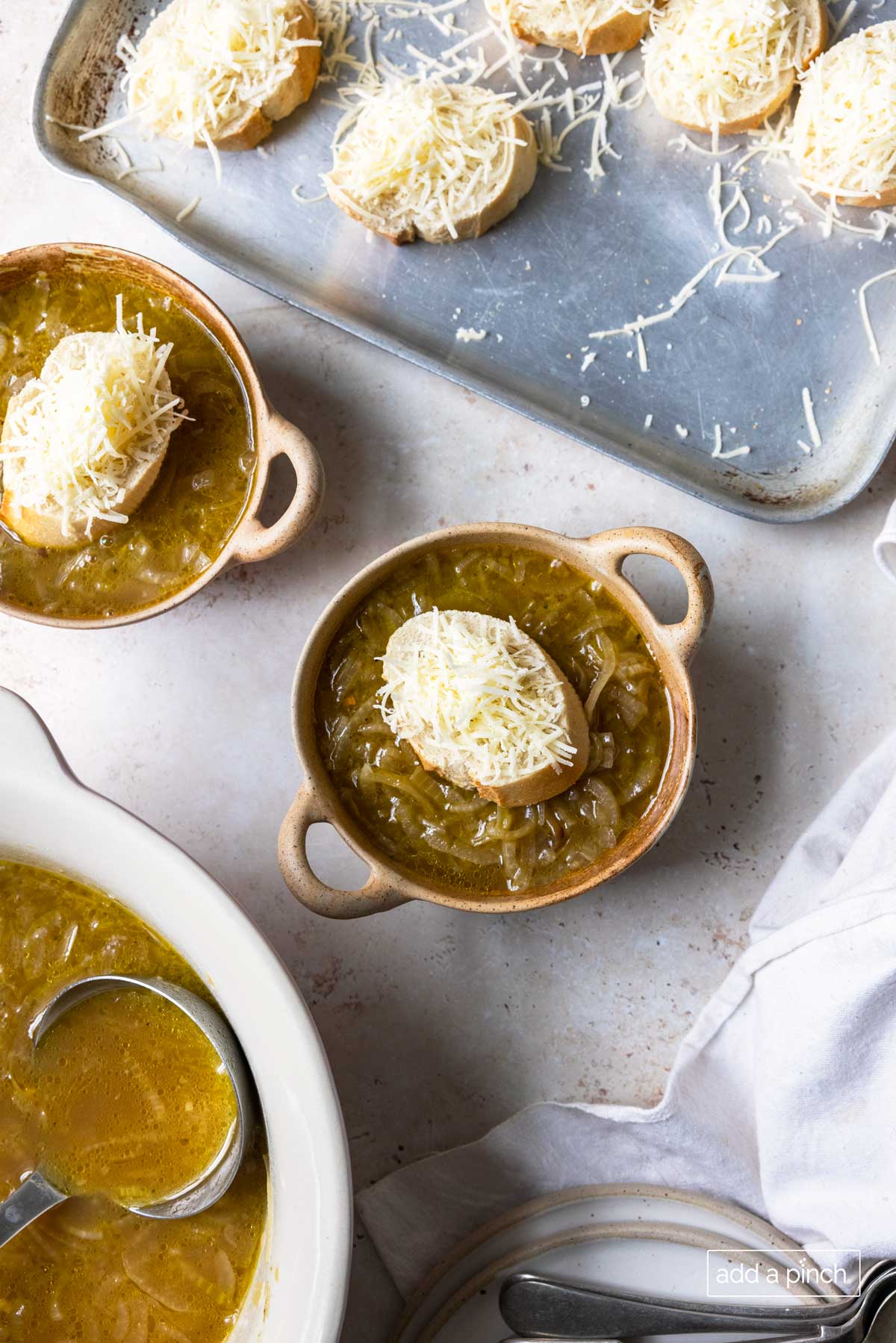 Baking pan has crusty french bread topped with grated cheese, along side crocks of soup topped with the cheese-topped bread ready to put in oven. 