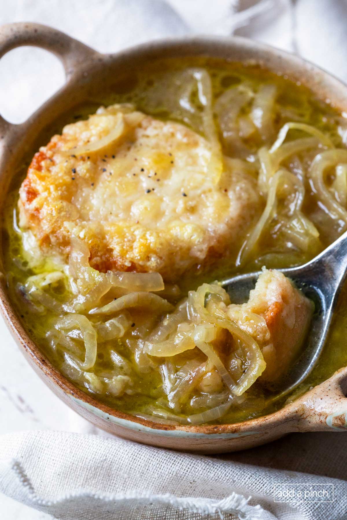 Stoneware bowl with french onion soup topped with cheesy crusty bread, being scooped by a spoon.