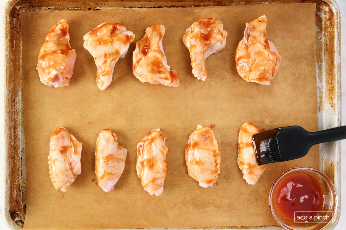 Chicken wings on a parchment lined baking sheet being brushed with bbq sauce.