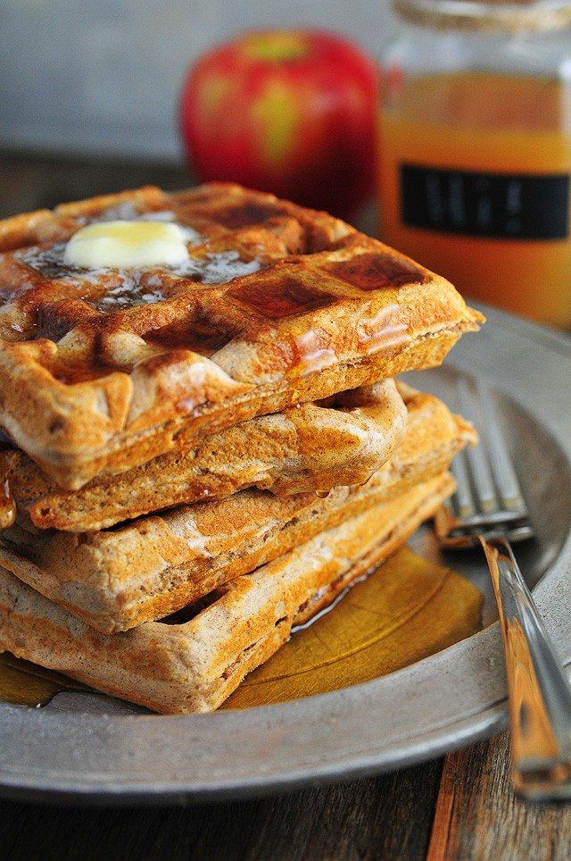 Apple-Cider-Waffles-DSC_0709 Stack of waffles on a pewter serving dish with fork. They are topped with melted butter and drizzled in maple syrup. Red apple and mug of apple cider in background // addapinch.com