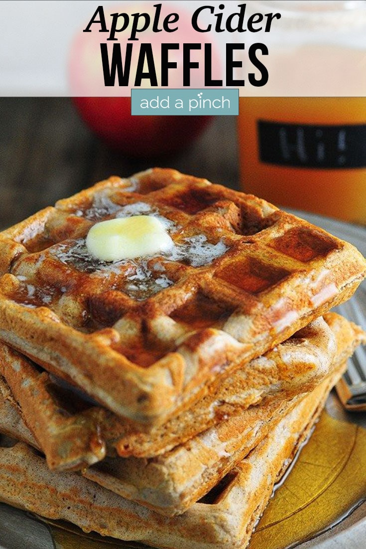 Stack of Apple Cider Waffles drizzled with melted butter and maple syrup on a pewter plate, with red apple and apple cider in background - with text - addapinch.com