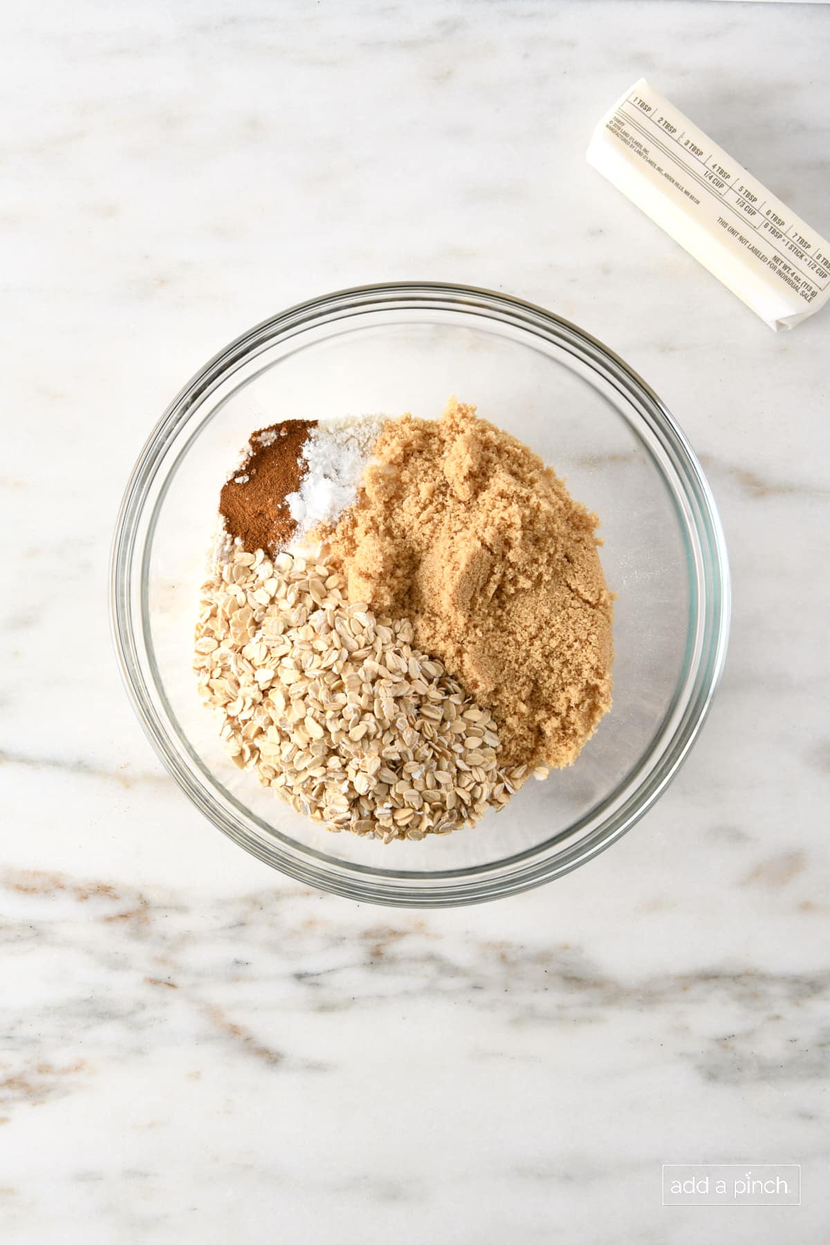 Glass bowl with oats, brown sugar, salt baking powder, cinnamon, with stick of butter to mix for oat streusel topping.
