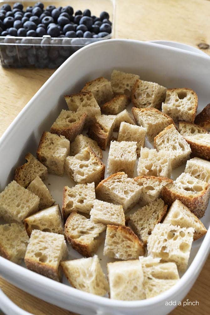 A white baking dish holds cubes of a rustic homemade bread. A container of fresh blueberries sits beside the baking dish.