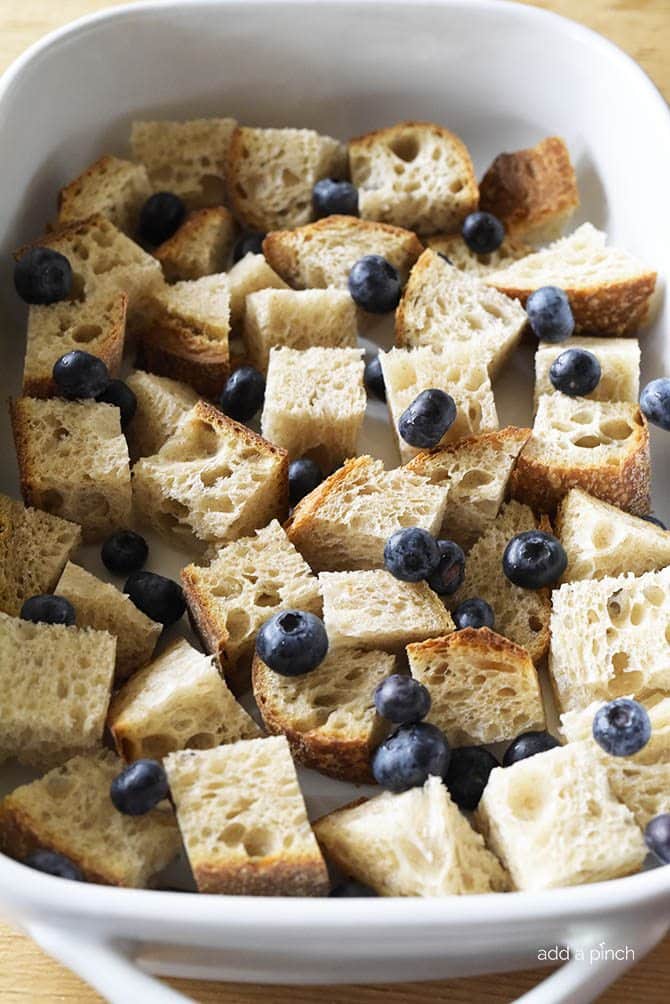 Liquid ingredients are poured over cubed sourdough bread and fresh blueberries in a white baking dish prior to baking. 