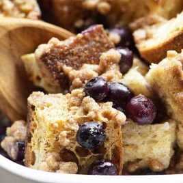 Baked Blueberry French Toast in a white baking dish is scooped with a wooden spoon, showing juicy blueberries and crisp streusel.