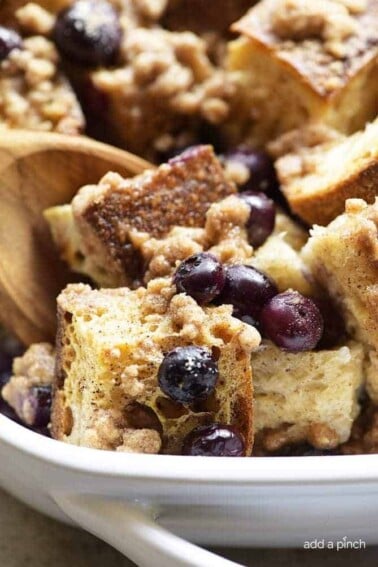 Baked Blueberry French Toast in a white baking dish is scooped with a wooden spoon, showing juicy blueberries and crisp streusel.