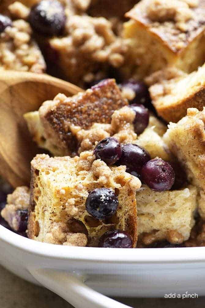 A wooden spoon scoops a serving of baked blueberry French toast casserole in a white casserole dish.