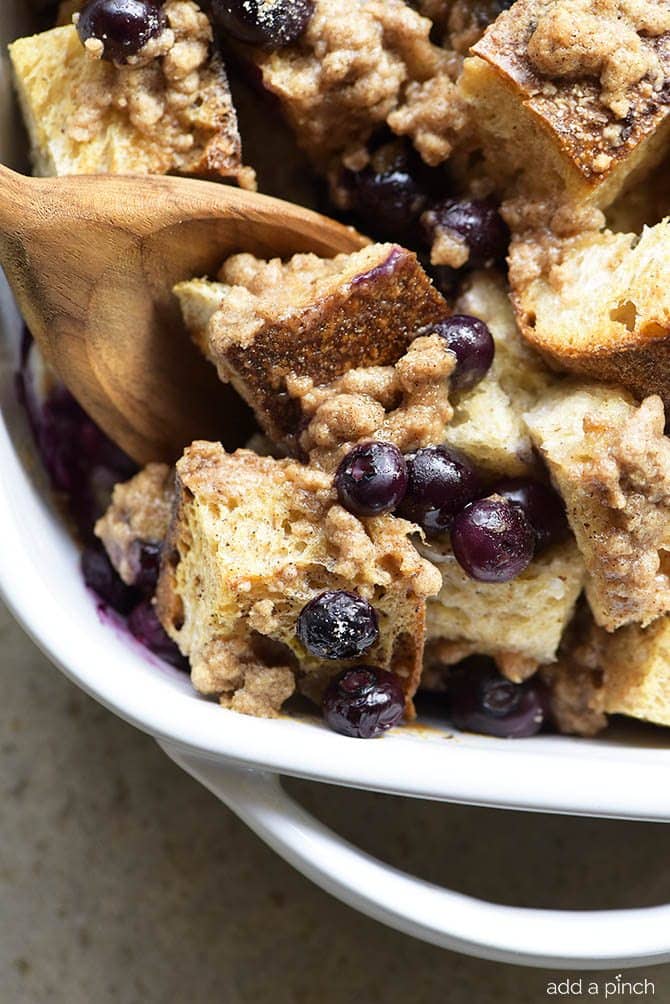 Closeup of wooden spoon scooping out a serving of baked blueberry French toast casserole. Shows juicy blueberries and streusel atop a creamy French toast. 