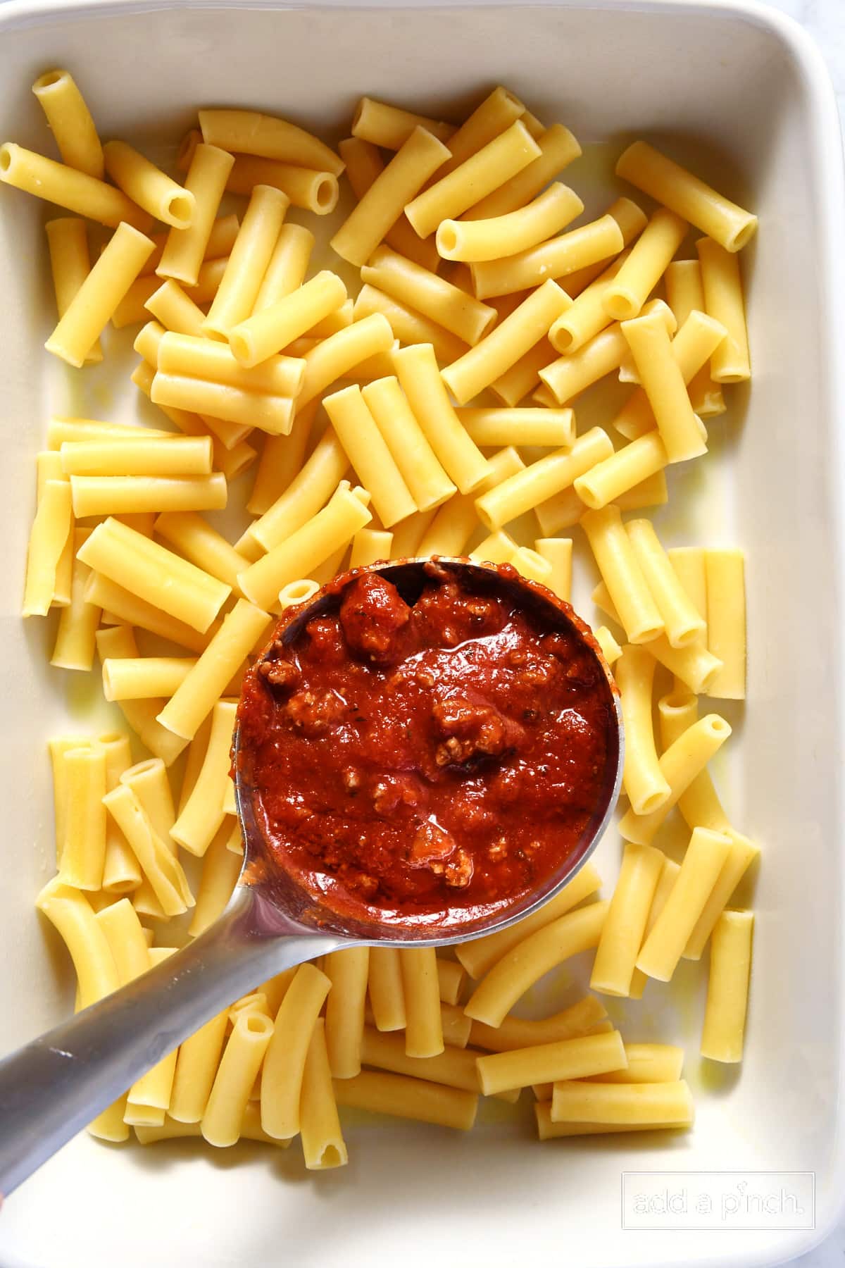 White baking dish filled with cooked noodles and a ladle of meat sauce.
