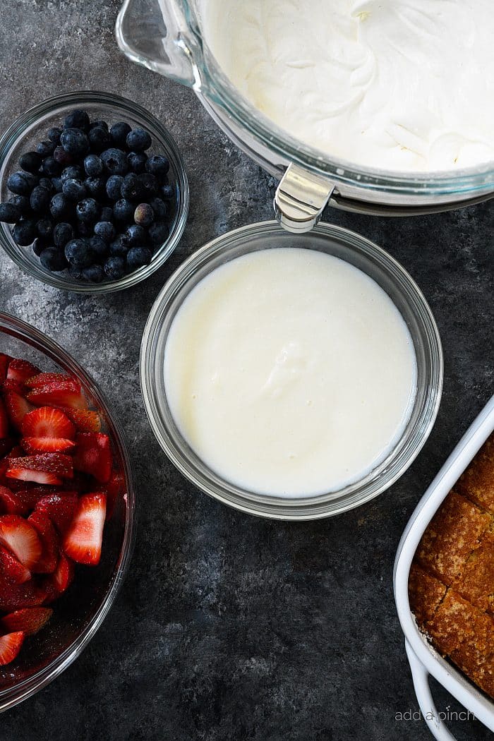 Ingredients for Berry Trifle, sliced strawberries, blueberries, whipped cream, vanilla pudding, vanilla cake cut into cubes, on a dark counter.