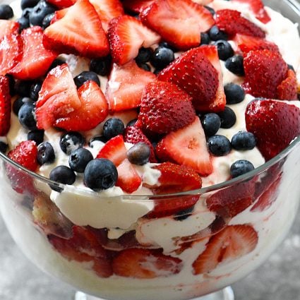 Glass trifle bowl holds a berry trifle dessert with layers of cream, vanilla cake, blueberries and strawberries.
