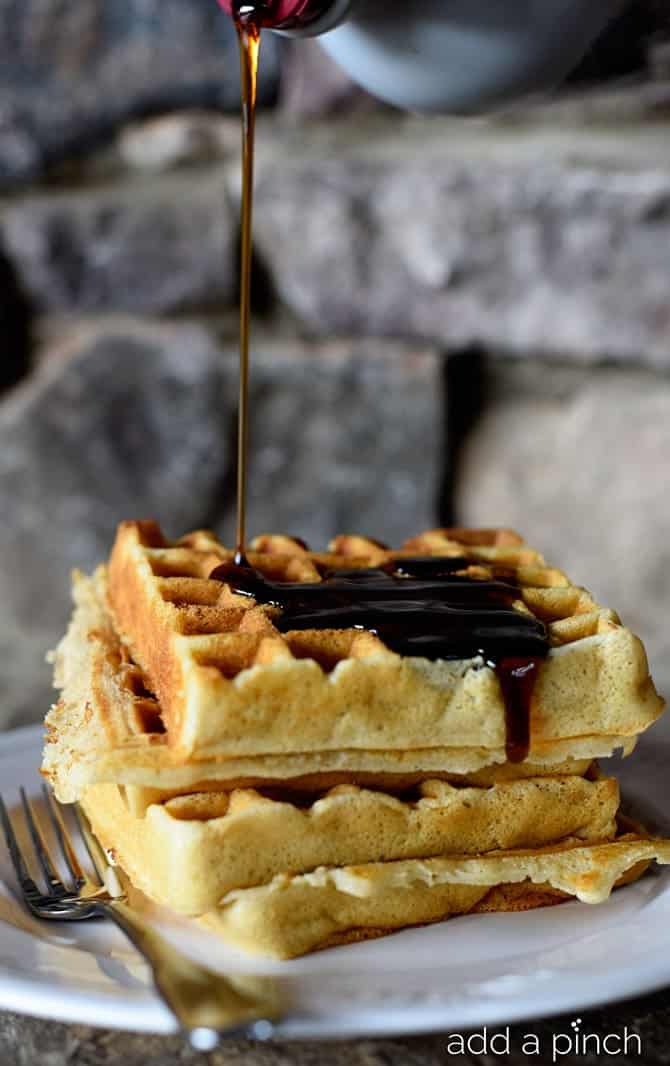 Our favorite buttermilk waffles stacked on a white plate with a fork and drizzled with a stream of syrup // addapinch.com