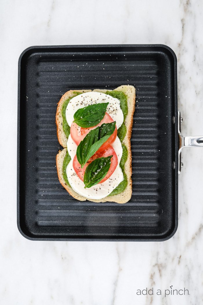 An assembled Caprese Grilled Cheese sandwich sits on grill pan showing layer of pesto, mozzarella, tomatoes and basil leaves // addapinch.com