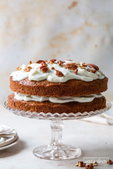 Carrot Cake recipe topped with cream cheese frosting, pecans on a clear glass cake stand.