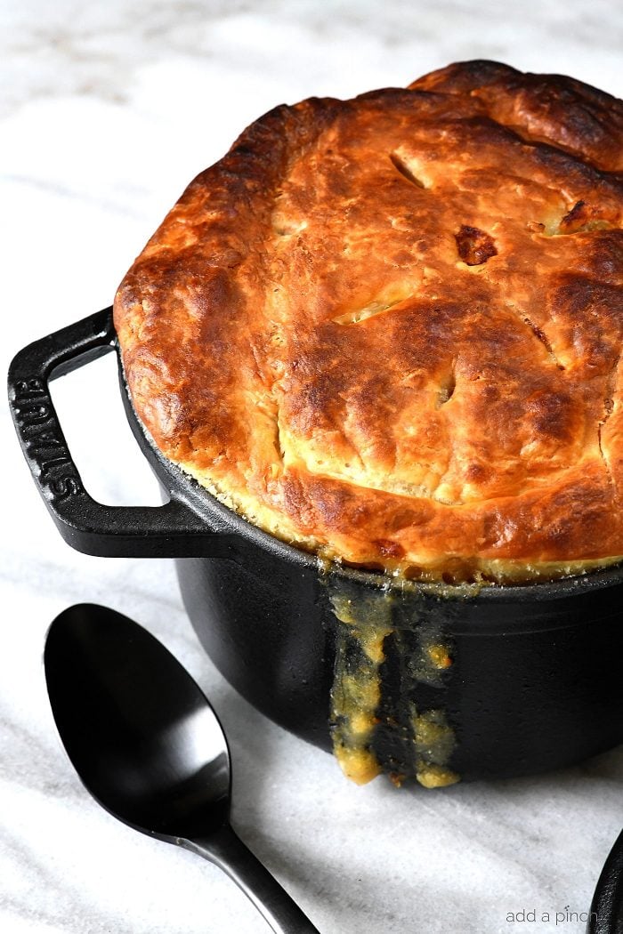 Black dutch oven is filled with chicken pot pie topped with a gold brown puff pastry top with thick juices running down the side. Sitting on marble countertop with black serving spoon. 