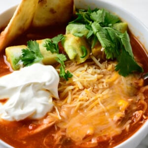 Bowl of chicken tortilla soup garnished with tortilla strips, avocados, sour cream, and shredded cheese, served in a white bowl.