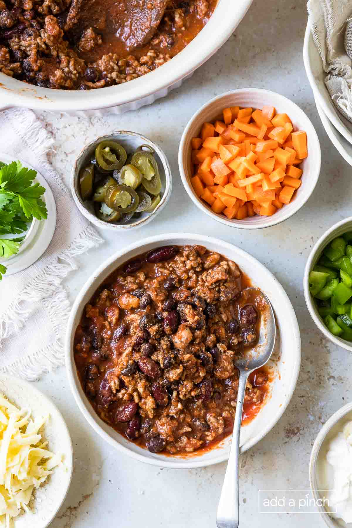Bowl of chili surrounded by bowls of optional toppings, such as cheeses, peppers, onions and more.  
