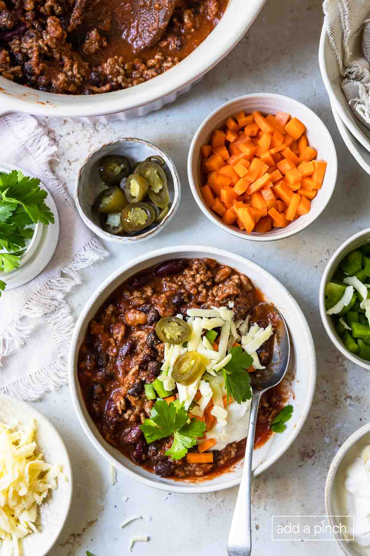 Bowl of chili garnished with herbs, jalapeno slices and cheese. It's surrounded by bowls of other ingredients for garnishing the chili.