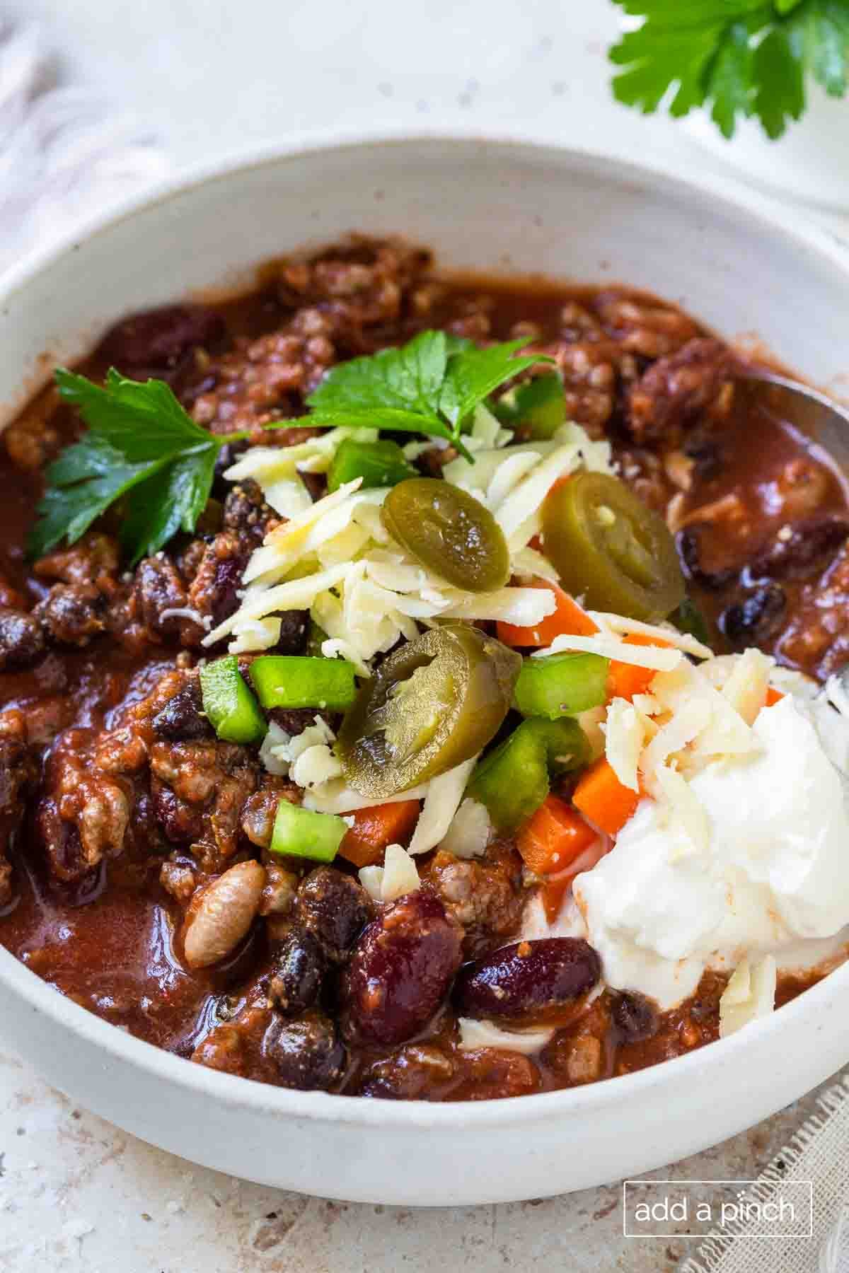 Bowl of chili with beef, beans, tomatoes and spices and then topped with sliced peppers, shredded cheese, sour cream and a sprig of fresh herbs.