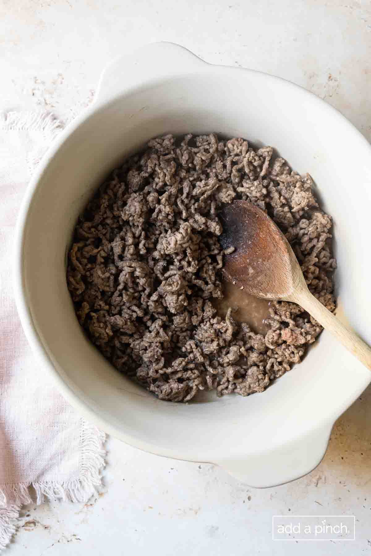 Browned ground beef in a large white Dutch oven with a wooden spoon. 