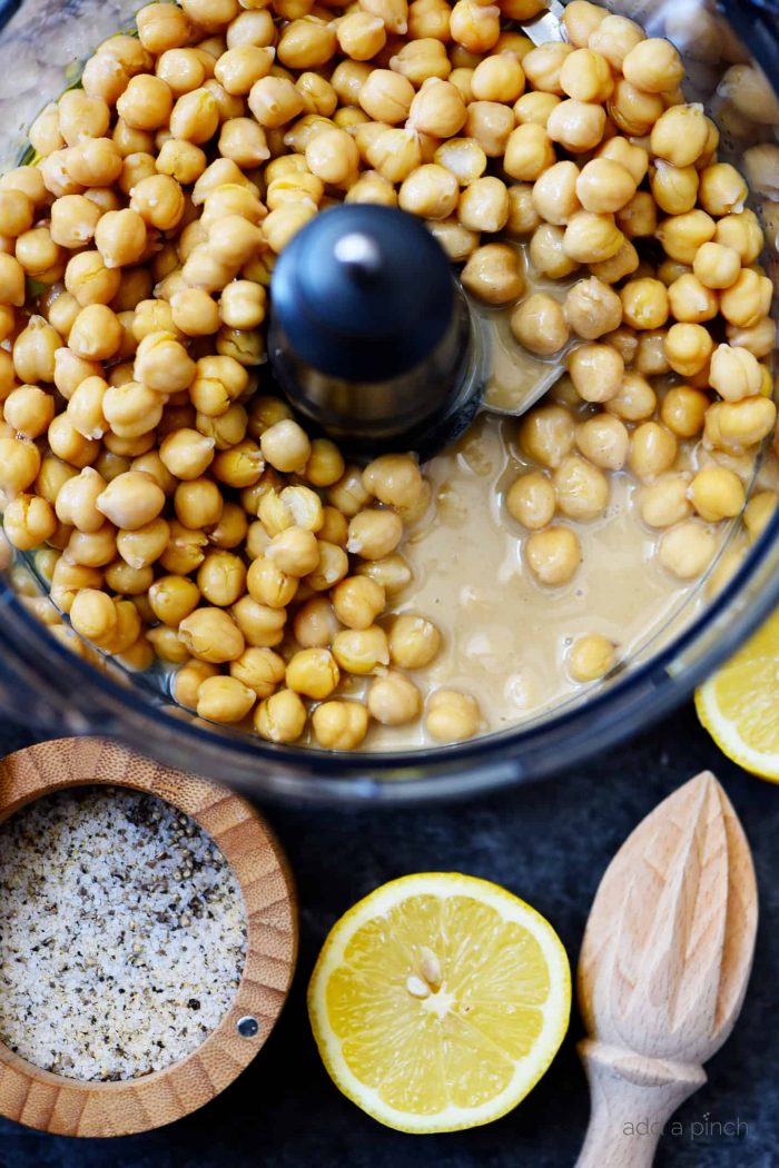Ingredients for hummus -chickpeas, tahini inside food processor container, surrounded by Stone House Seasoning, sliced lemon and wooden lemon juicer // addapinch.com
