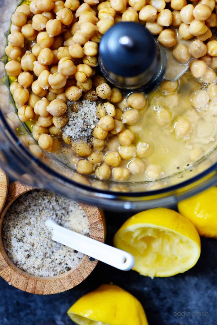 Hummus ingredients in food processor container surrounded by lemon rinds and Stone House seasoning with a measuring spoon  // addapinch.com