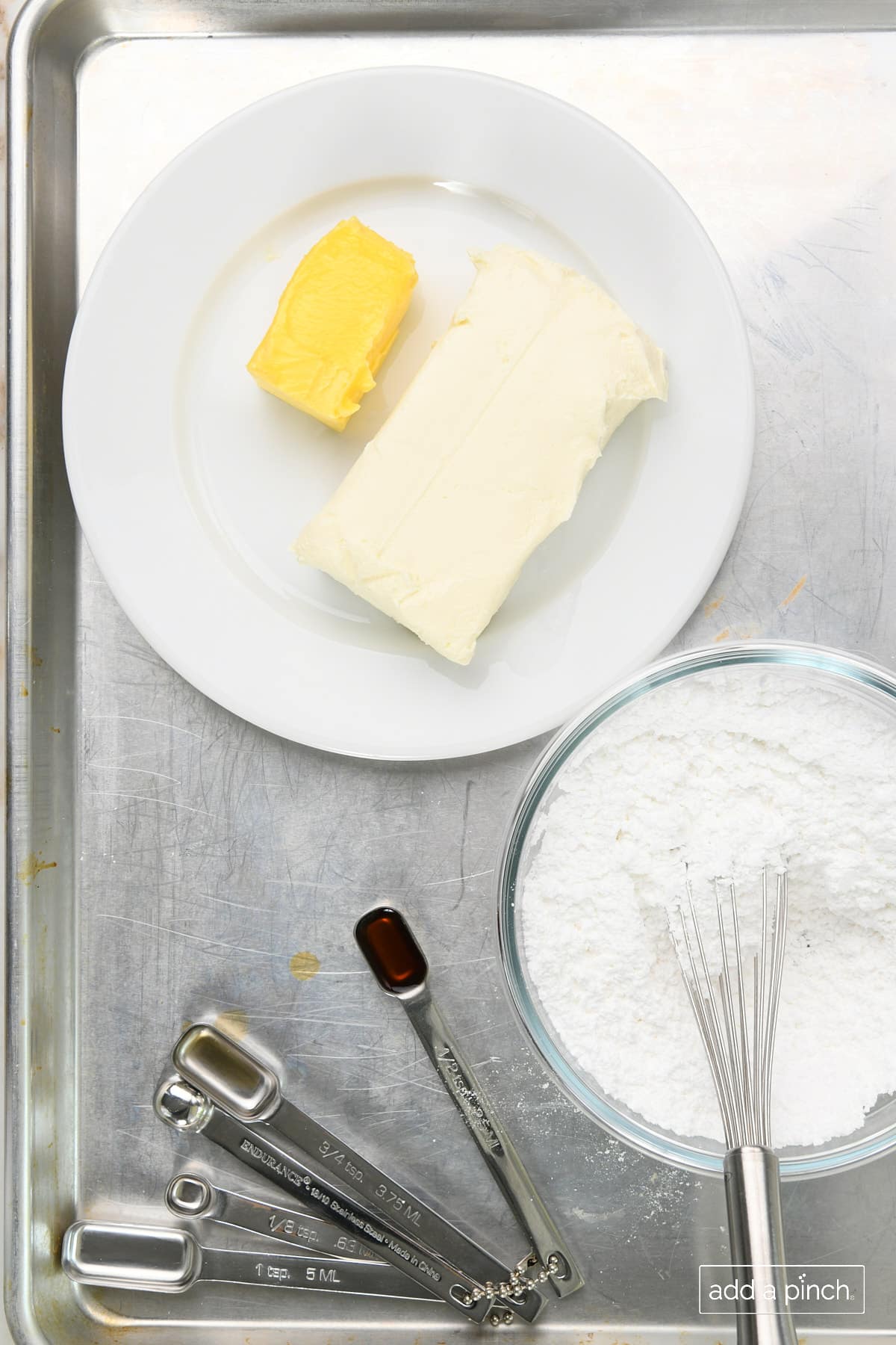 Ingredients needed to make the best cream cheese frosting are measured into bowls and measuring spoons and on baking sheet along with softened cream cheese and butter.