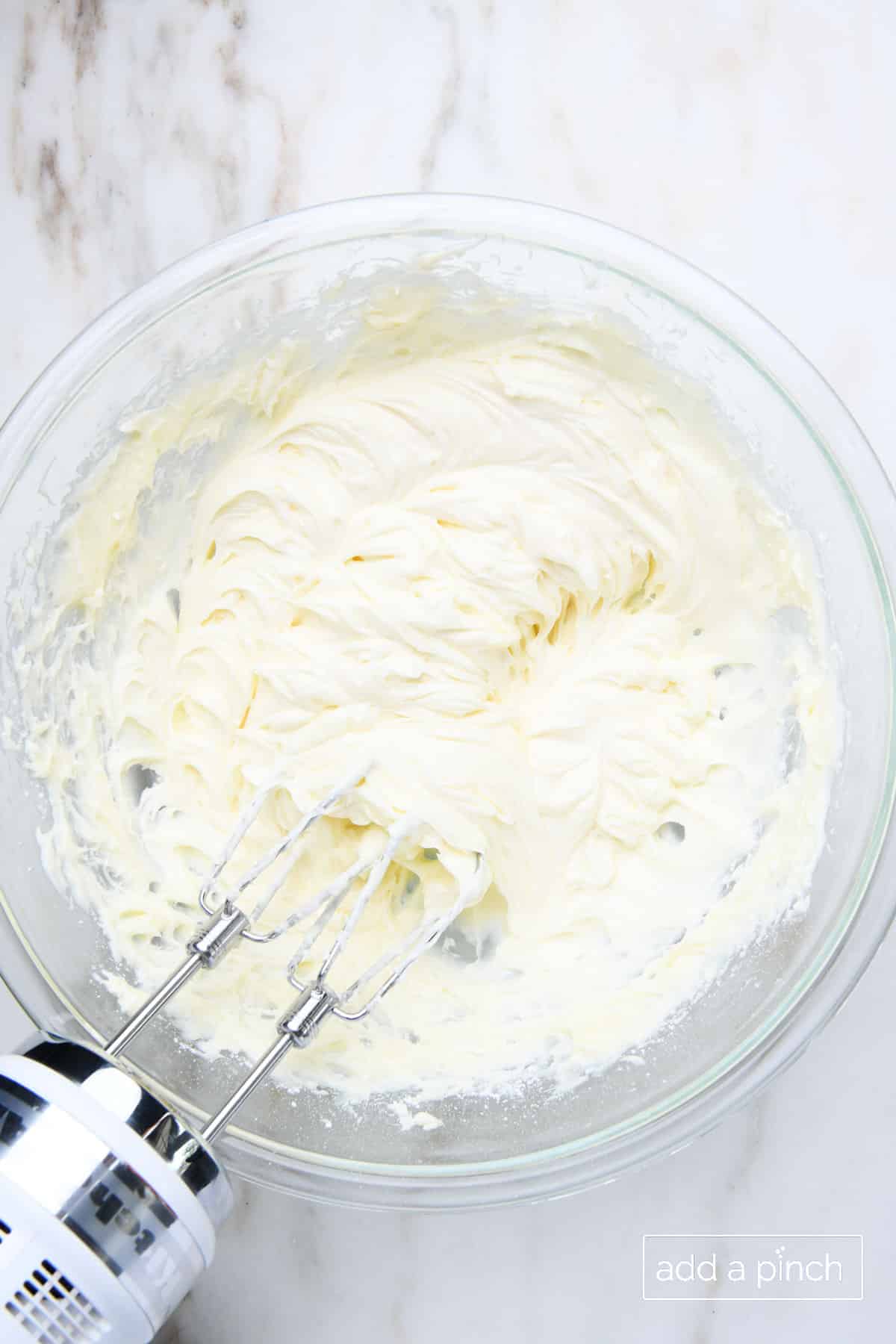 Overhead of glass mixing bowl with fluffy cream cheese frosting mixed with a hand mixer. Bowl is sitting on marble countertop. 