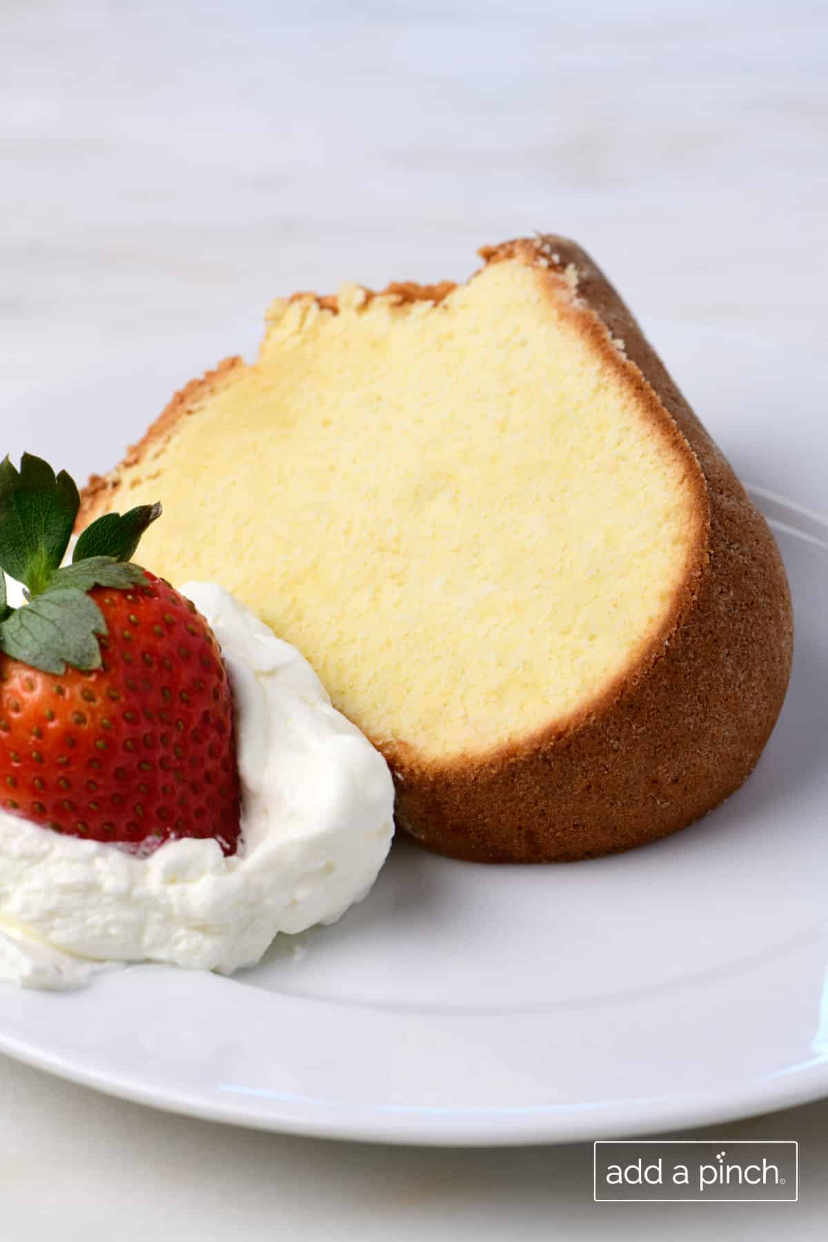 Slice of cream cheese pound cake with dollop of homemade whipped cream and red strawberry on a white dessert plate.