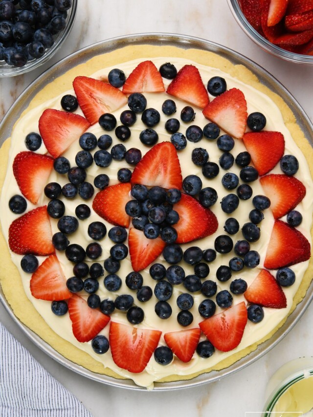 Fruit pizza on a marble surface surrounded by bowls of blueberries, strawberries, and cream cheese frosting.