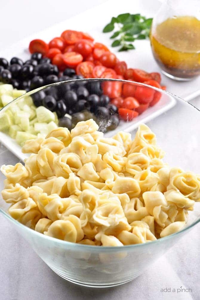 Cooked tortellini in glass bowl with chopped tomatoes, cucumbers and ripe black olives on white dish. Italian dressing in glass dispenser  // addapinch.com