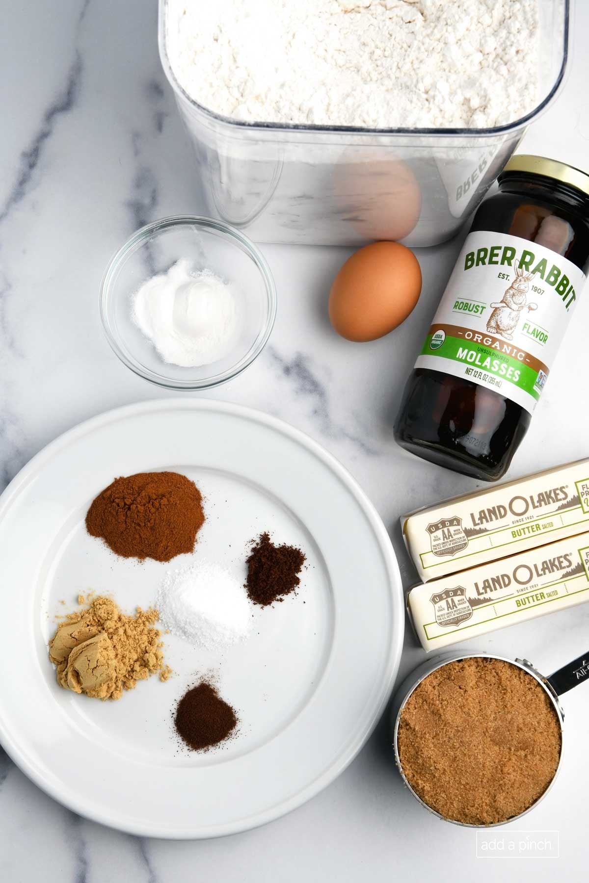 Ingredients to make gingerbread cookies are laid on a marble counter: butter, brown sugar, molasses, salt, cinnamon, ginger, cloves, vanilla extract, egg, baking powder, baking soda, espresso powder, and flour.