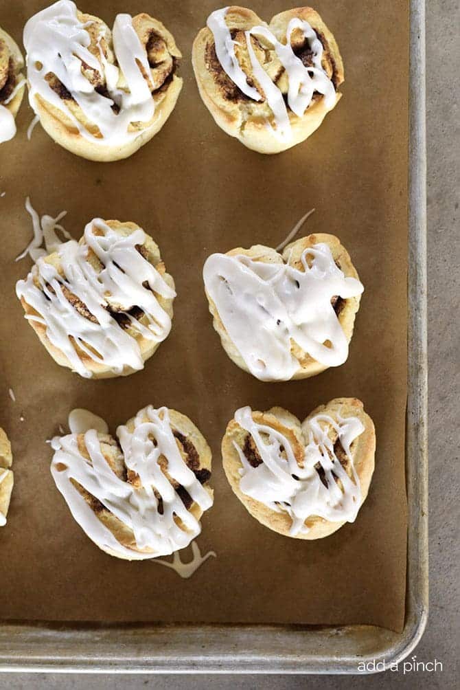 Multiple iced cinnamon roll biscuit hearts on a parchment lined baking sheet. 