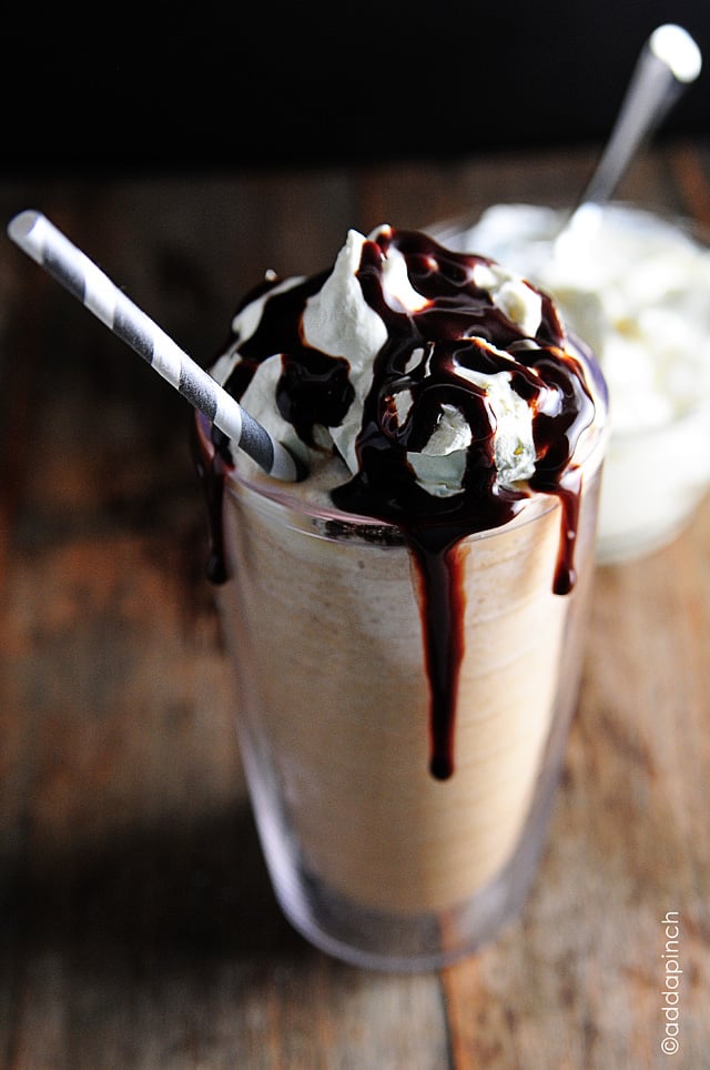 Homemade-Frappuccino-DSC_0560 Frosty glass holds a cold Frappuccino coffee drink topped with homemade whipped cream, drizzled with chocolate syrup that is running down the sides of the glass. A straw is inserted in the glass for drinking. Bowl of whipped cream sits in background with a spoon in it.