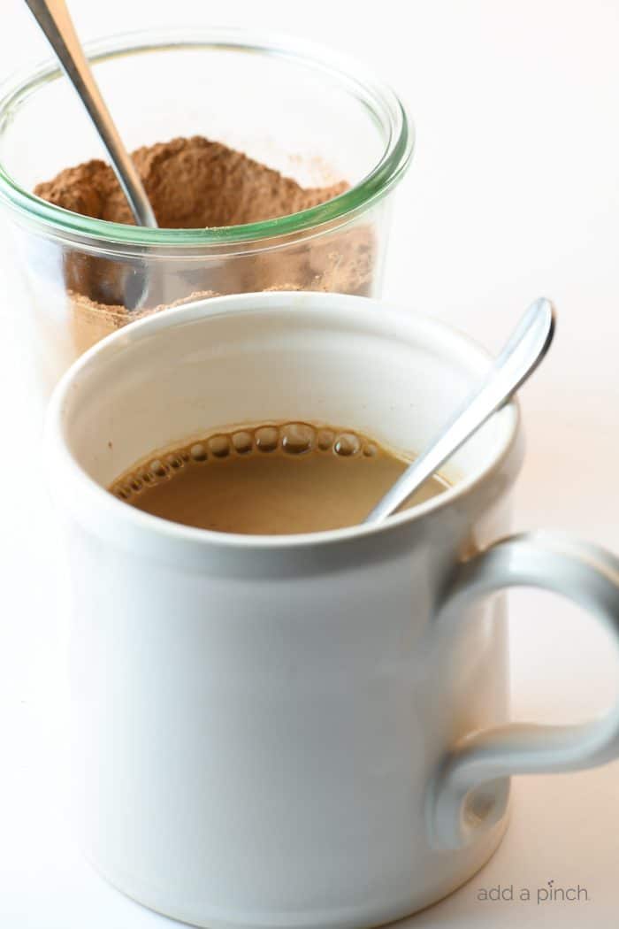 Photograph of white mug filled with hot cocoa and a clear jar of homemade mix behind it on a white counter.