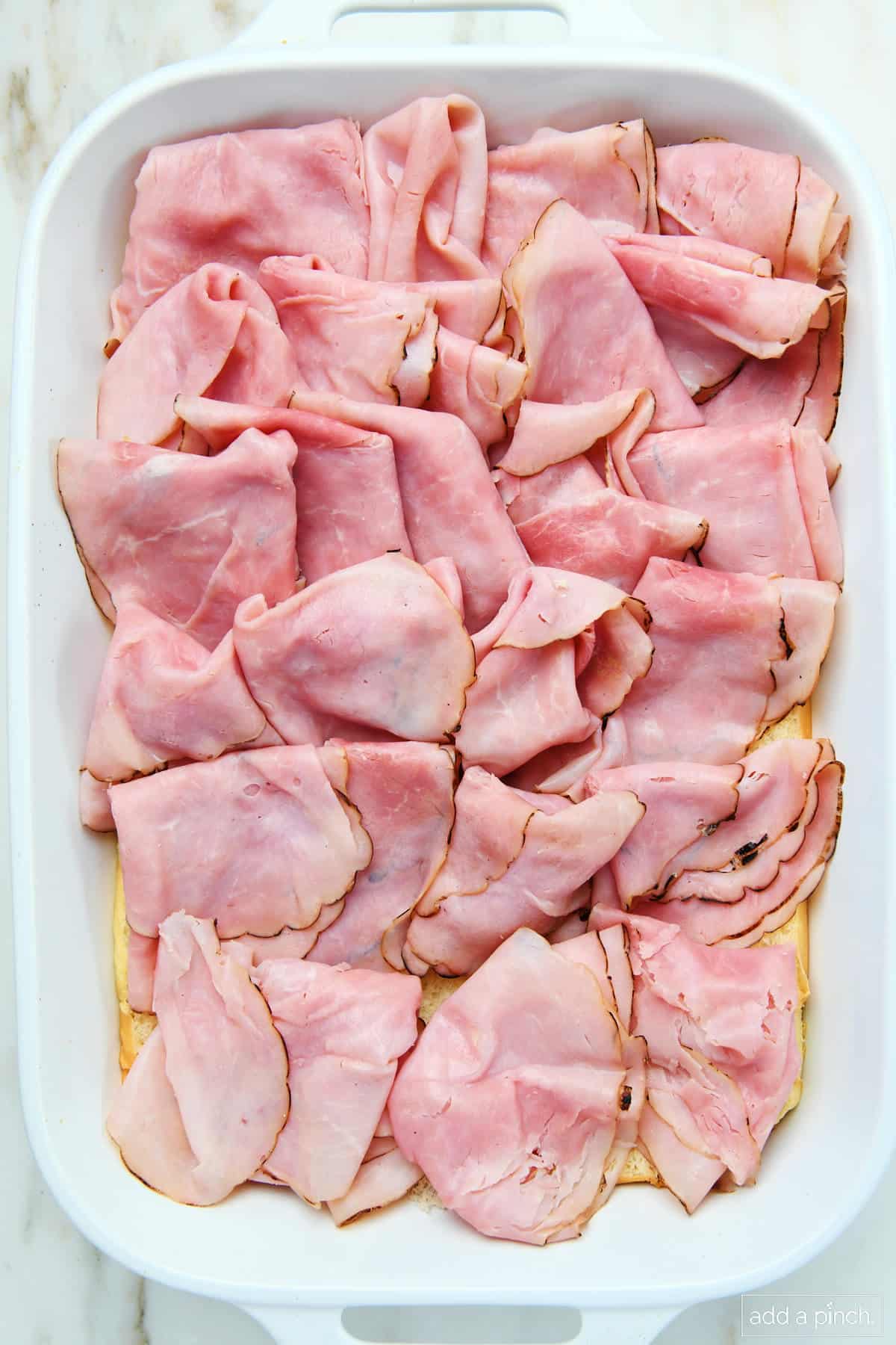 Ham layered onto bottom half of soft rolls in a baking dish.