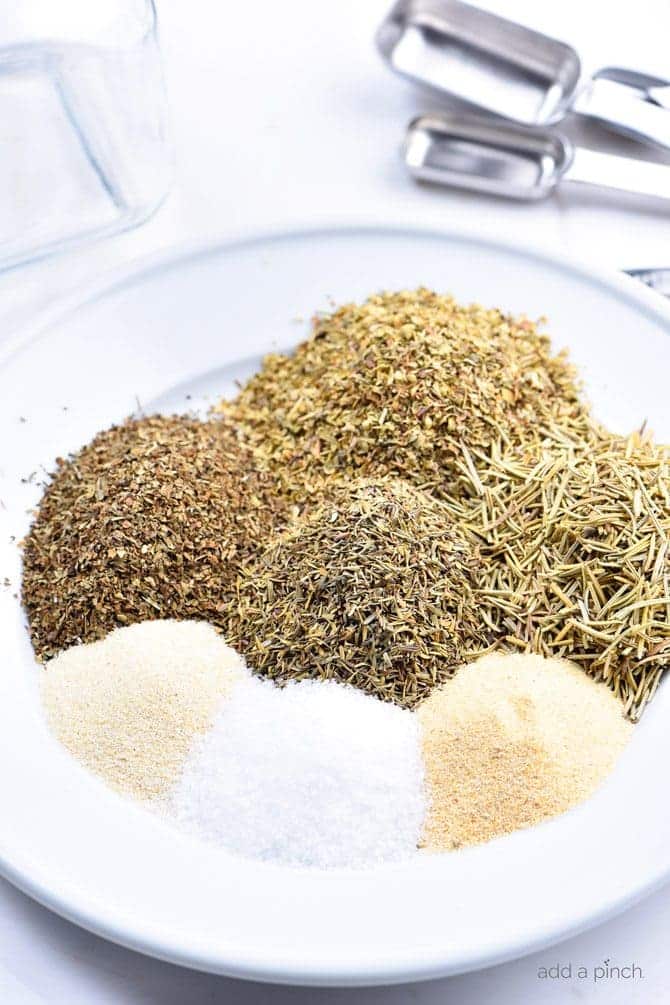 Measuring spoons, glass container surround dried herbs and spices on a white plate waiting to mix to make Italian Seasoning Mix. 