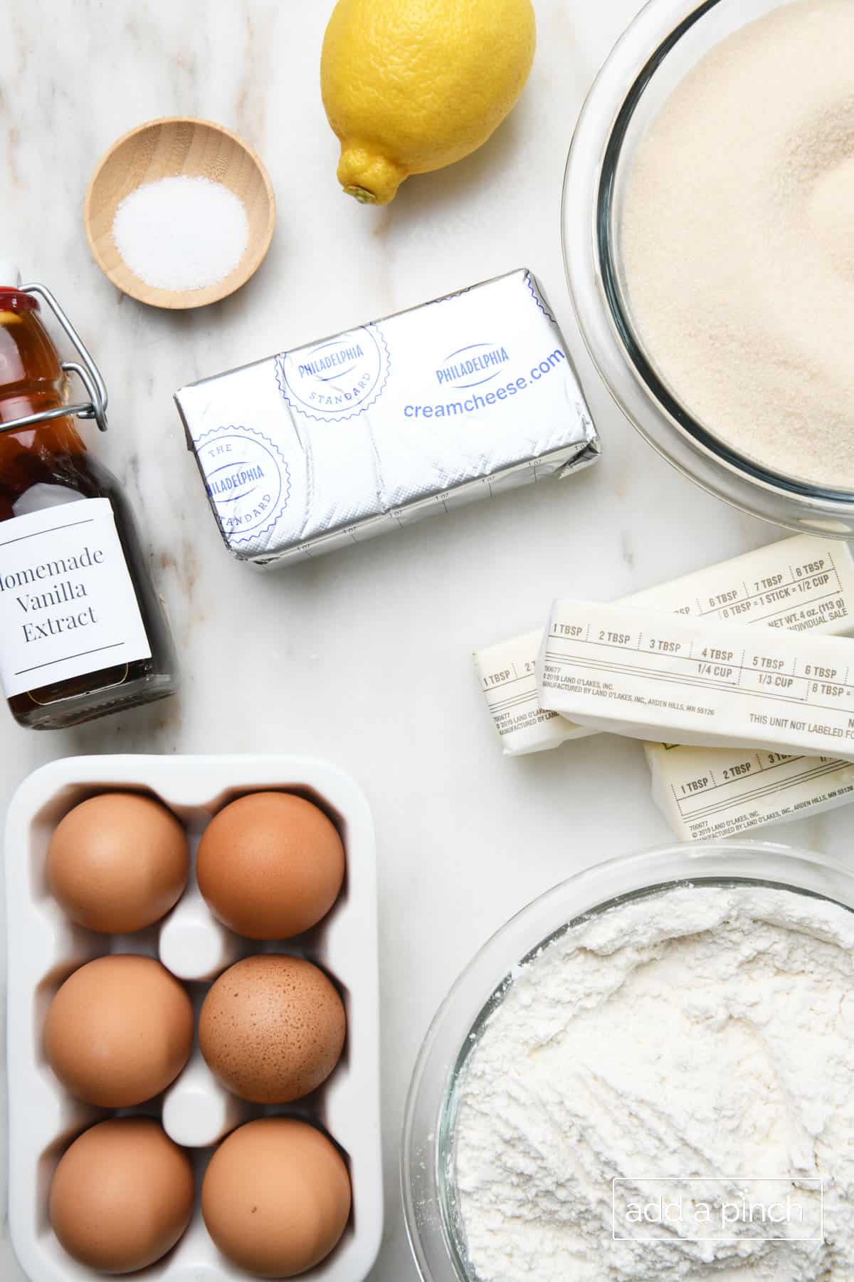Ingredients for Lemon Pound cake lie on a marble countertop - fresh lemon, sugar, sticks of butter, flour, eggs, homemade vanilla extract, cream cheese and salt.