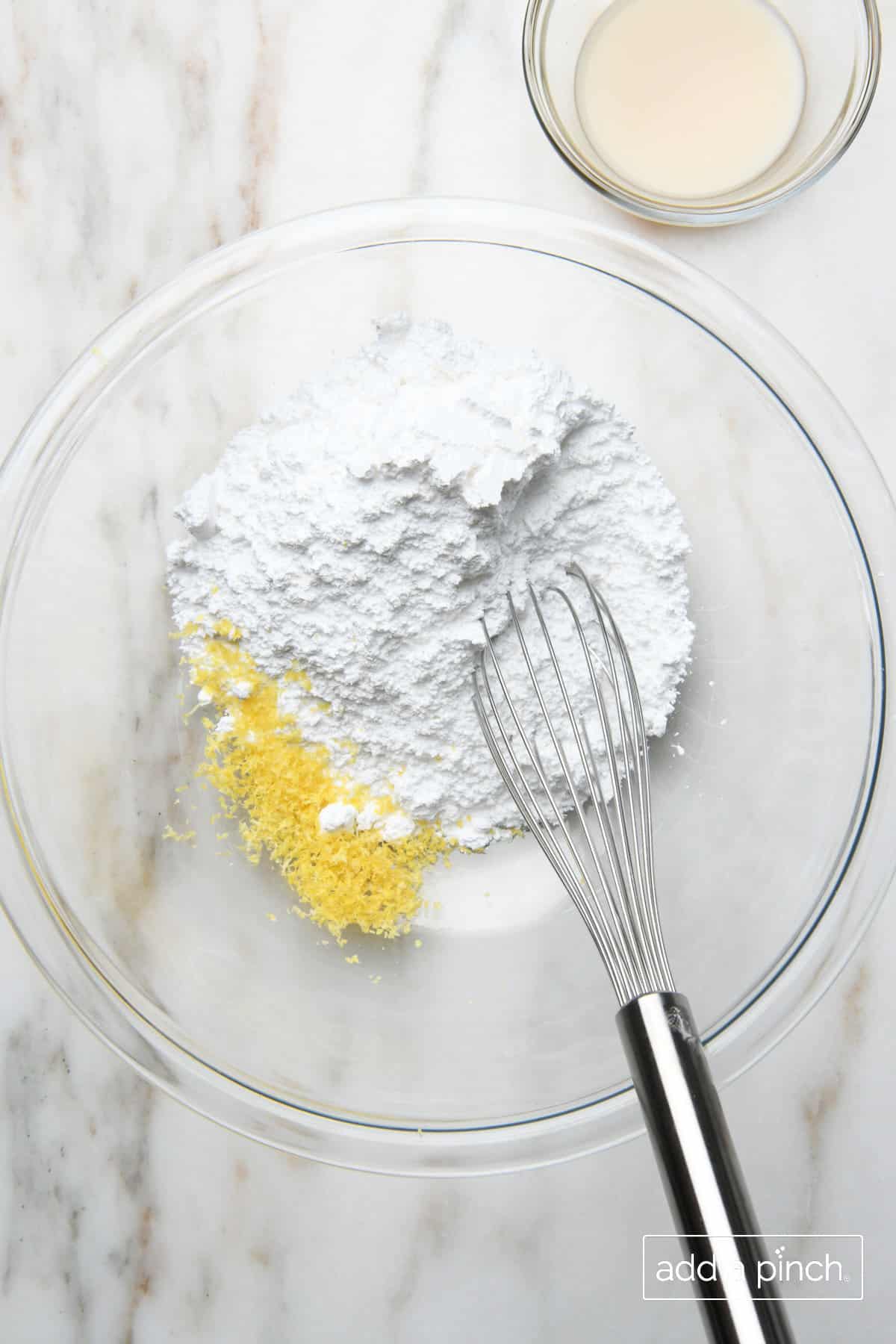 Ingredients for the Lemon Buttermilk Glaze sit in a glass mixing bowl with a whisk on a marble countertop. A small bowl with buttermilk sits beside the larger mixing bowl.