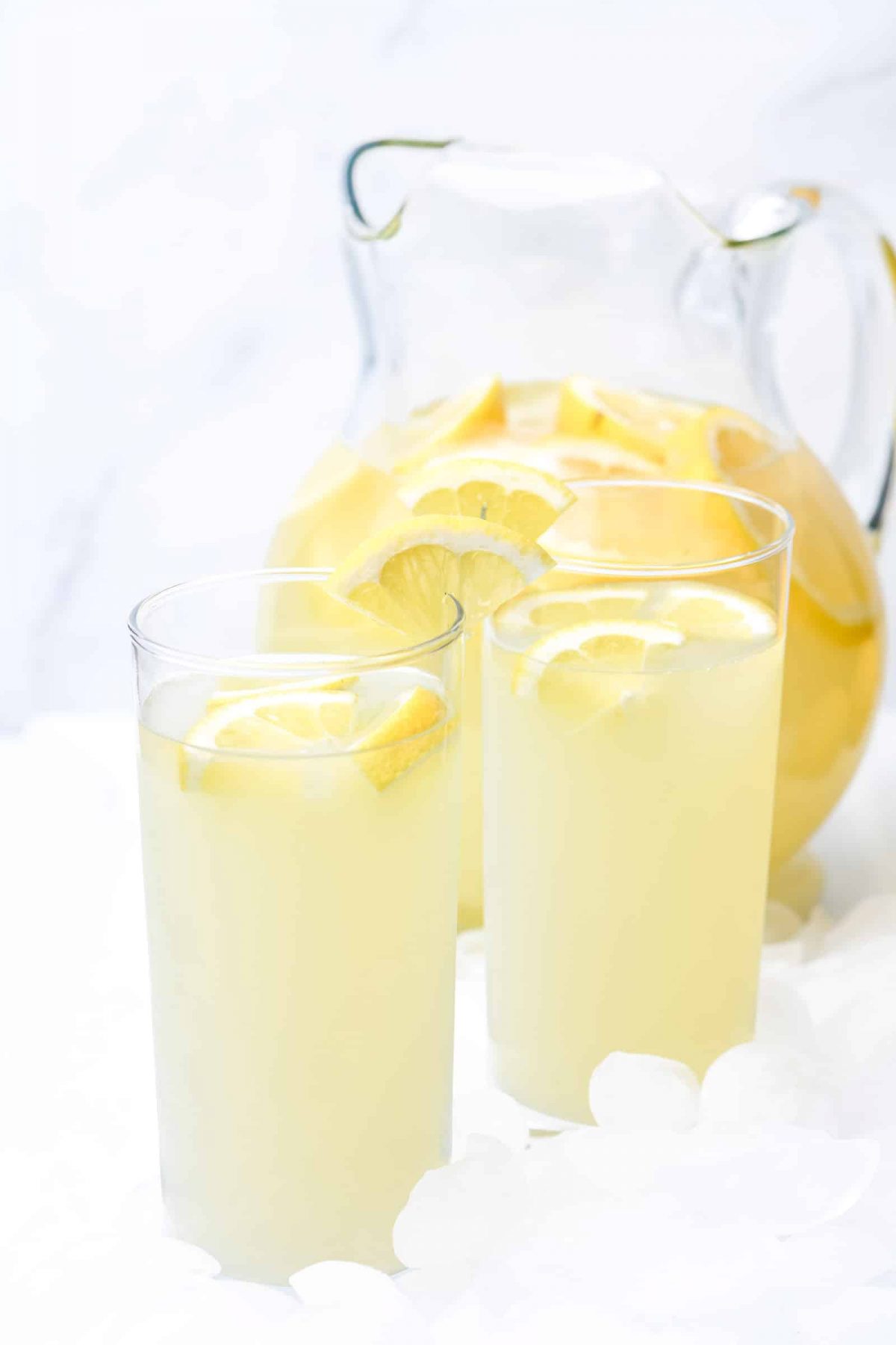Photo of a clear pitcher of lemonade with lemon slices and 2 clear glasses of lemonade with lemon slices on a white background.