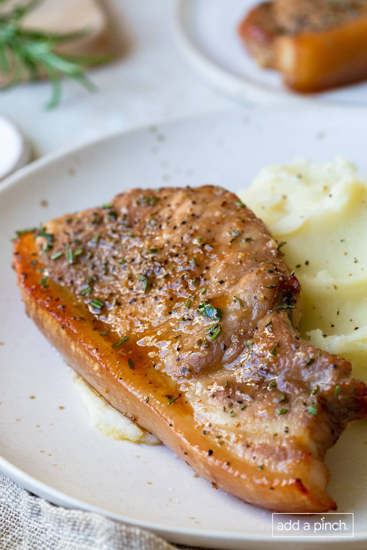 Pork chop on a white plate with mashed potatoes on a white surface. Background of fresh herbs and more meat.
