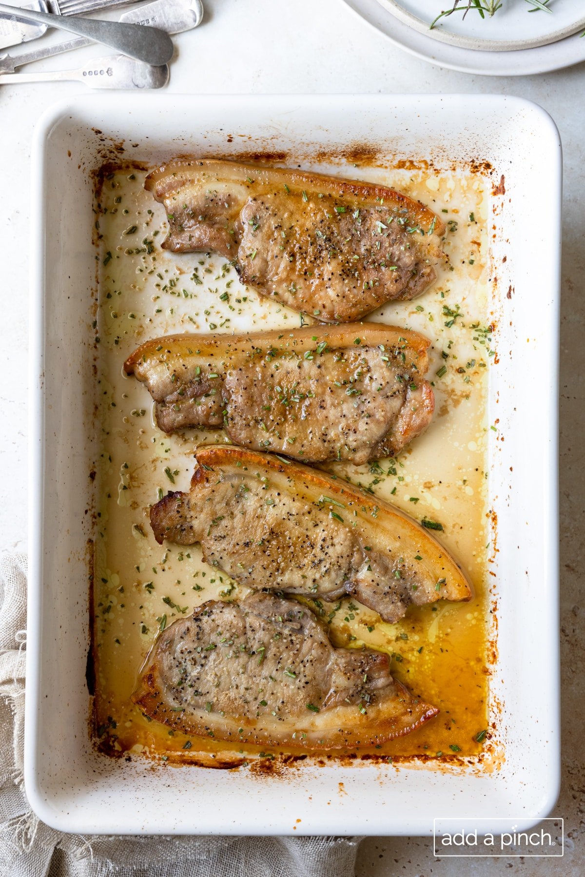 Photo of baked pork chops in a white baking dish with a stack of plates in the background on a white surface.
