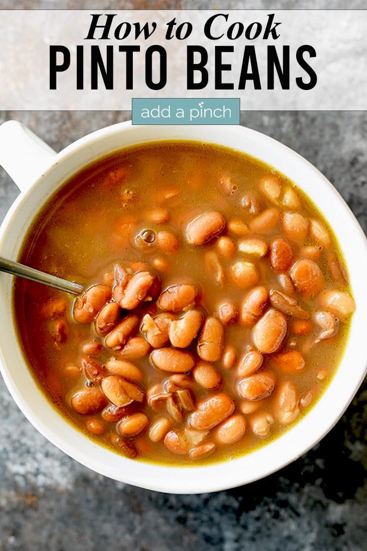 Cooked bowl of pinto beans in a white bowl with spoon on a dark counter.