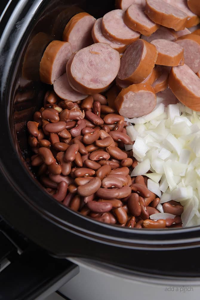 Slow cooker insert filled with red beans, chopped onion, sliced sausage.