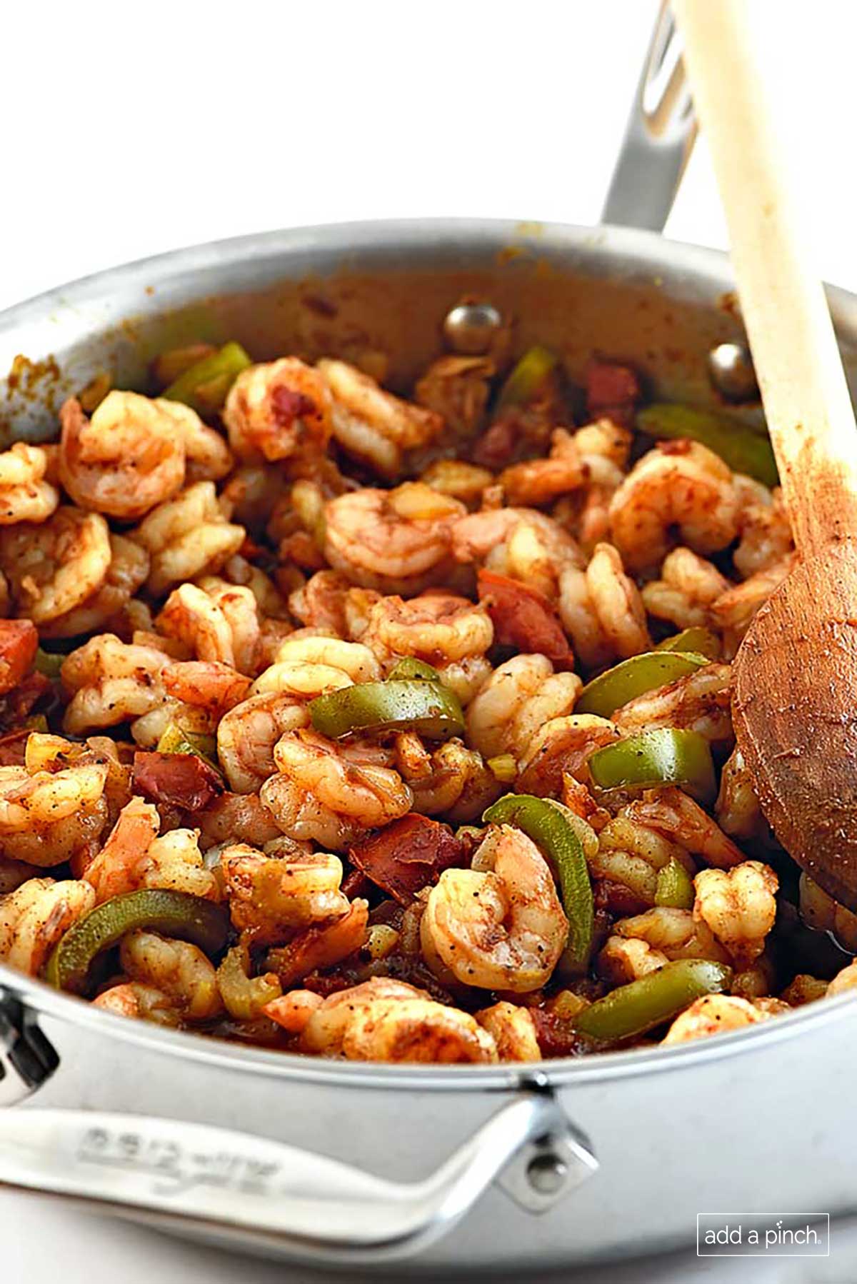 Photo of Shrimp Creole with shrimp, green pepper slices and tomatoes with a wooden spoon in a stainless steel skillet.