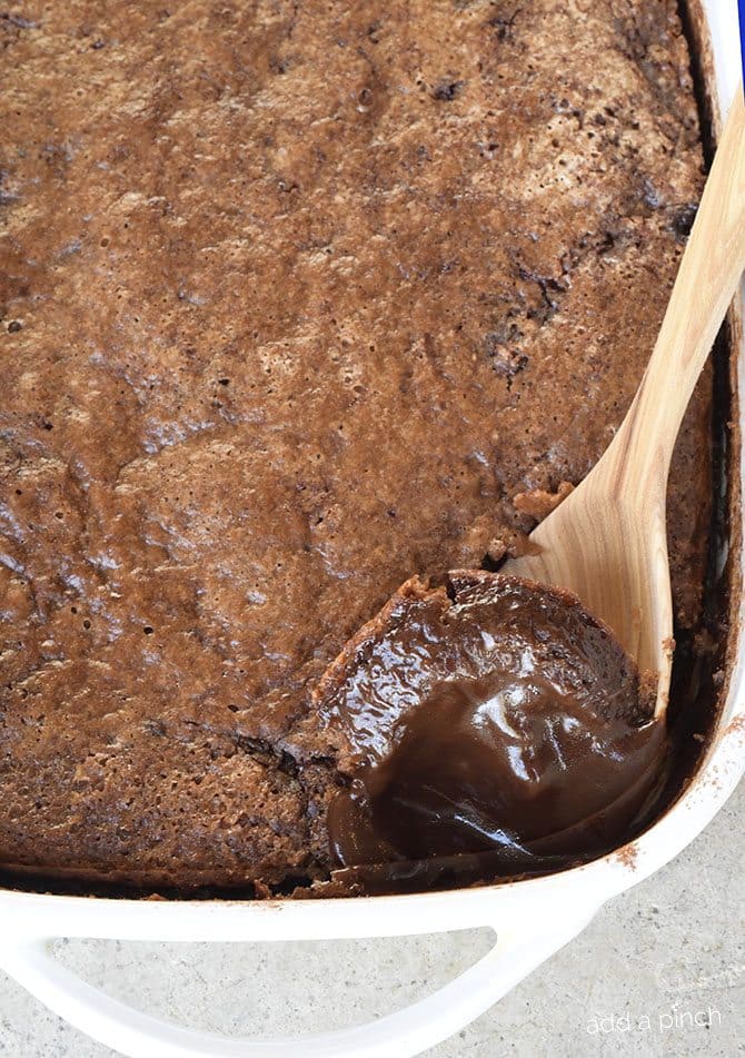 White baking dish and wooden spoon scooping into brownie-like top to reveal fudge inside of chocolate cobbler.