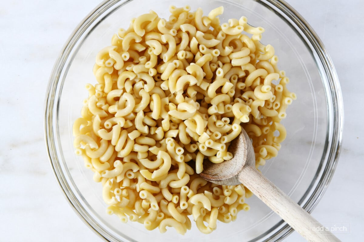 Glass bowl and wooden spoon, with cooked and drained elbow macaroni noodles.