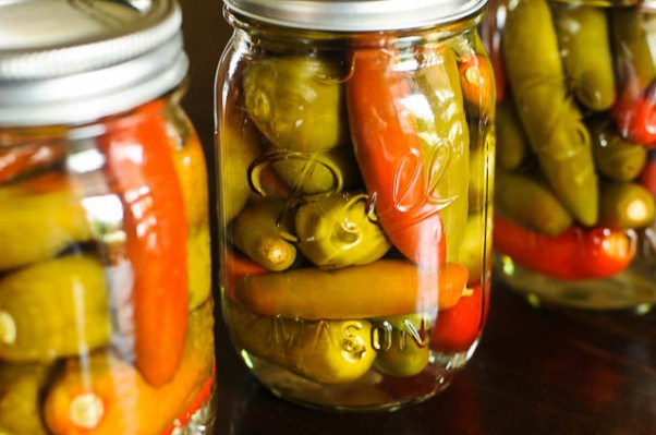 Glass jars hold pepper sauce with colorful peppers inside. 