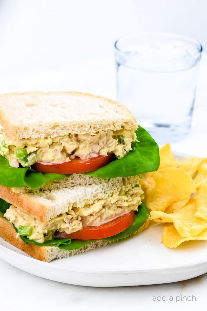 White plate with tuna salad sandwich on white bread. Has sliced tomatoes and bibb lettuce leaves, side of chips and a glass of water.  // addapinch.com